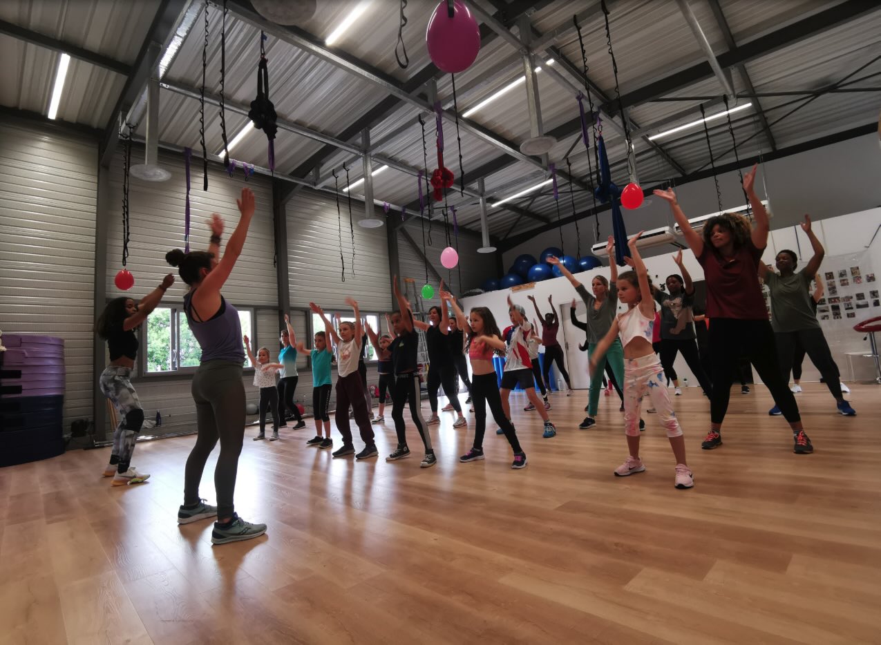 Cours collectif intergénérationnel dans une salle de sport avec enfants et adultes suivant une coach sur musique, ambiance conviviale et dynamique